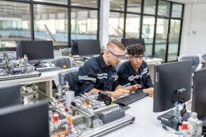 two quality engineers sitting a desk covered in electronics, looking at a computer screen