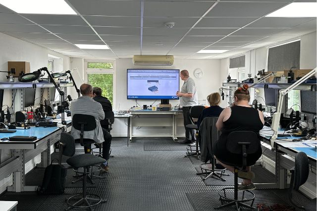 a master IPC trainer standing at the front of an electronics workshop showing a presentation to 4 students sitting at work benches