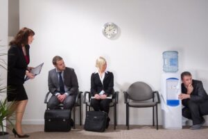 a room of candidates waiting for interviews and one of the candidates is hiding behind the water cooler because he is nervous for his interview