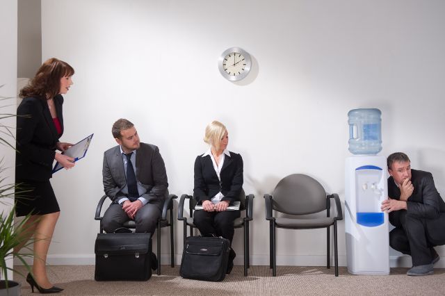 a room of candidates waiting for interviews and one of the candidates is hiding behind the water cooler because he is nervous for his interview
